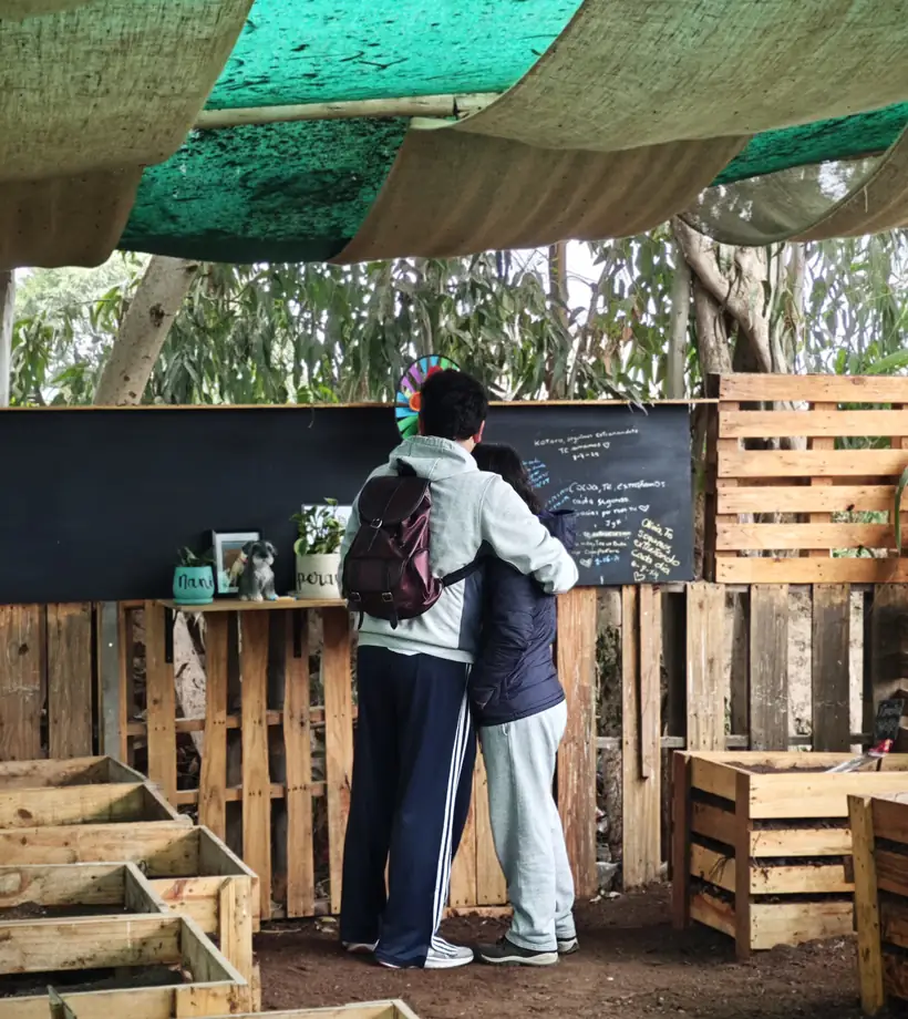 Dos personas abrazadas frente al muro de mensajes del camposanto, entre las cunas composteras de madera
